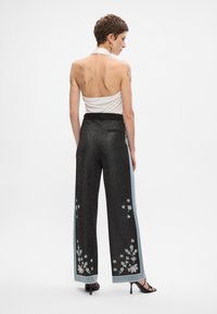 Pantalons large noirs et scintillants avec des rayures latérales bleues et de la broderie florale sur le bas des jambes. Modèle portant un haut halter blanc.