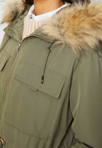 Olivově zelená parka bunda s kapucí lemovanou kožešinou, se zapínáním na zip a dvěma předními kapsami s klopami. Hladká textura a nastavitelný stahovací šňůrka.