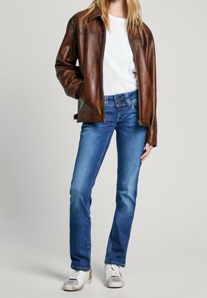 Personne portant une veste en cuir marron sur une chemise blanche, un jean bleu avec une fermeture à double bouton, et des baskets blanches avec des accents beige.