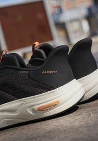 Schwarze und graue Sportschuhe mit weißen gepolsterten Sohlen mit der Aufschrift "CLOUDFOAM" und "RAPIDFIT", auf einer Betonoberfläche platziert.