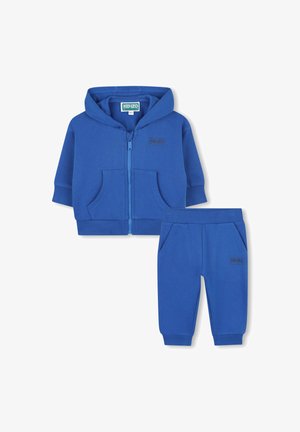 Haut à capuche zippé bleu avec poches avant et pantalon assorti. Les deux pièces présentent une texture douce et un logo discret sur le tissu.