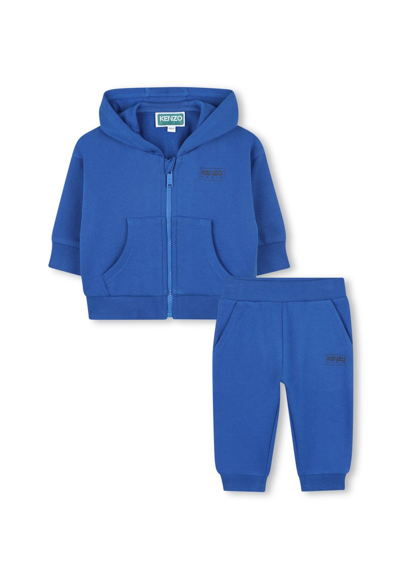 Haut à capuche zippé bleu avec poches avant et pantalon assorti. Les deux pièces présentent une texture douce et un logo discret sur le tissu.