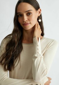 Maglietta beige a maniche lunghe con scollo a onda, dettagli con bottoni ai polsini e una texture liscia e morbida. Gli orecchini hanno forma di cuore.