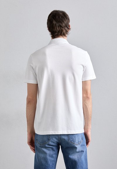 T-shirt en coton blanc avec un col rond simple et des manches courtes, associé à un jean bleu, vu de dos contre un fond clair.