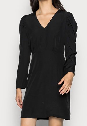 Frau trägt ein schwarzes Mini-Kleid mit langen Ärmeln, gepufften Schultern und V-Ausschnitt, steht vor einem schlichten Hintergrund.