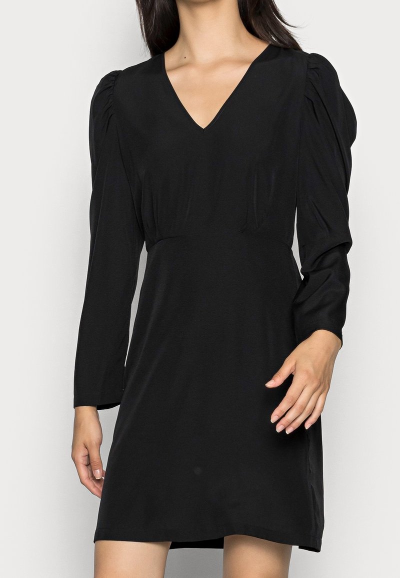 Femme portant une mini-robe noire à manches longues avec épaules bouffantes et col en V, debout devant un fond uni.