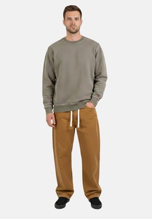 Man staat naar voren gericht, draagt een grijze trui met ronde hals, bruine broek met trekkoord en zwarte sneakers, met één hand in de zak.