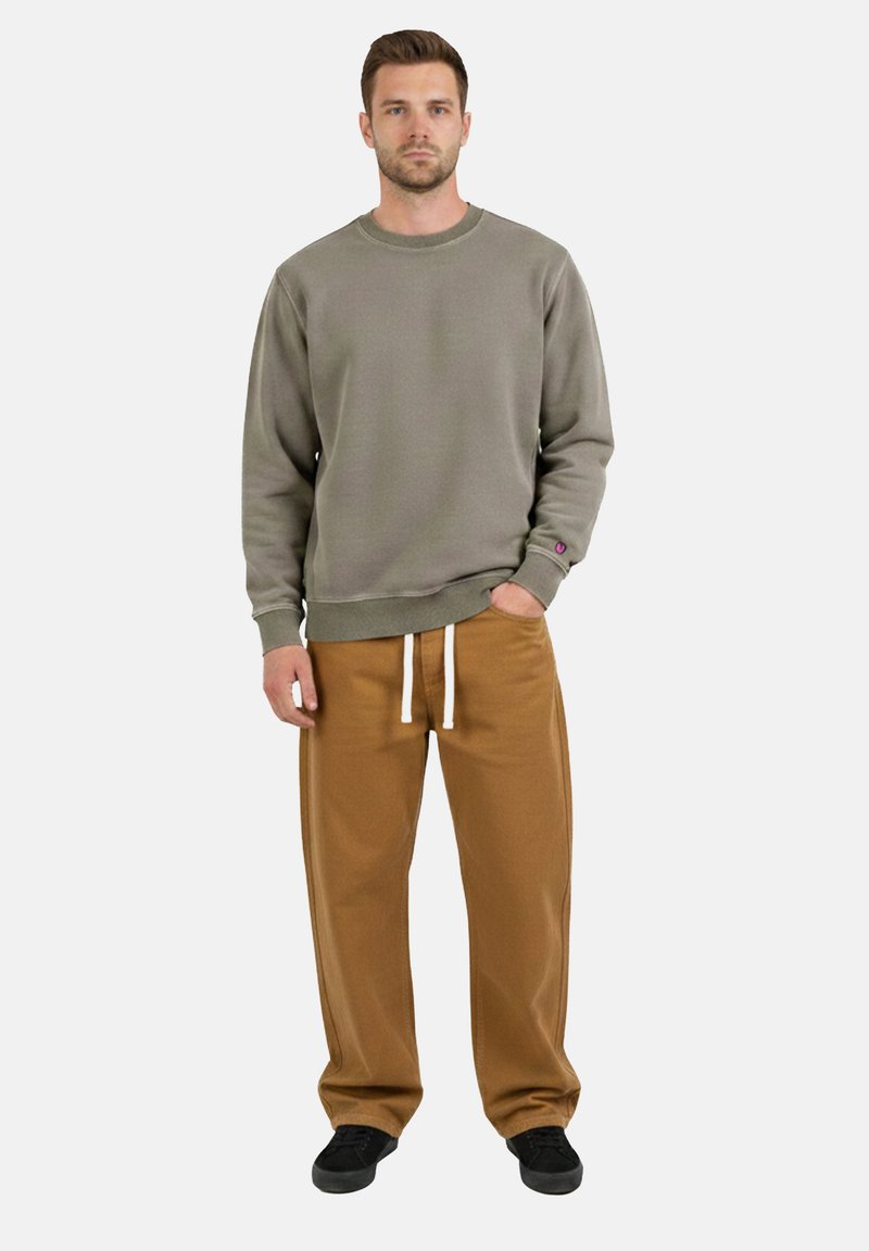 Homme debout face à l'avant portant un sweat-shirt gris à col rond, un pantalon marron à cordon de serrage et des baskets noires, avec une main dans la poche.