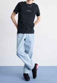 T-shirt nera a maniche corte con logo "JORDAN", jeans a vestibilità ampia di colore blu chiaro e sneakers nere con accenti rossi. Sfondo semplice.