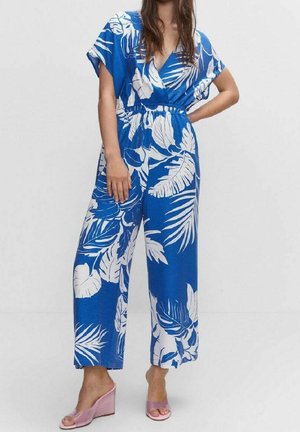 Woman wearing a blue jumpsuit with white tropical leaf patterns and pink open-toe wedge sandals, standing with one hand on her hip.