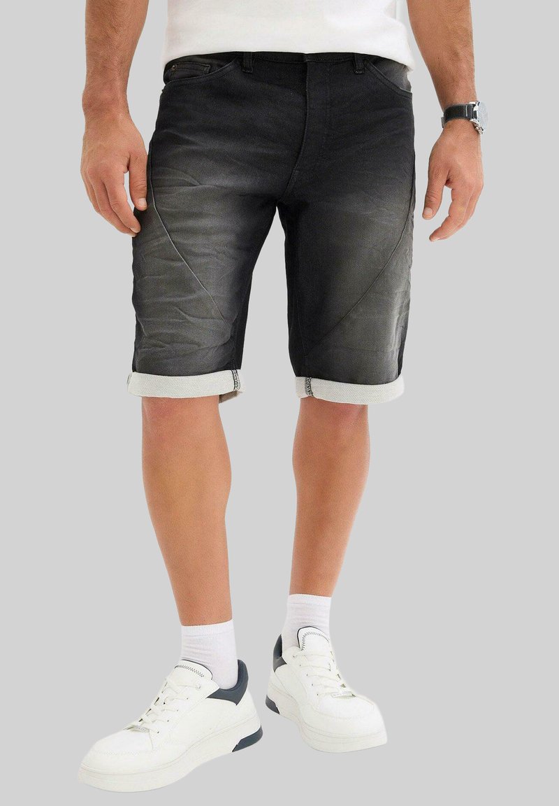Shorts en denim noir avec un ourlet replié, présentant une texture lisse et des détails de coutures diagonales subtils. Assortis avec des baskets blanches et des chaussettes.