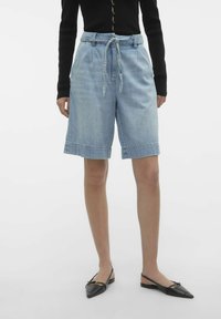 Shorts de mezclilla azul claro con cintura ajustable con cordón, bolsillos laterales y dobladillo enrollado. Combinados con zapatos planos negros de punta.