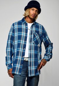 Camisa de cuadros azul de botones con mangas largas, que presenta tonos de azul y marrón, un bolsillo en el pecho y un corte holgado. Usada sobre una camiseta blanca.