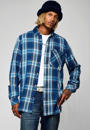 Camisa de cuadros azul de botones con mangas largas, que presenta tonos de azul y marrón, un bolsillo en el pecho y un corte holgado. Usada sobre una camiseta blanca.