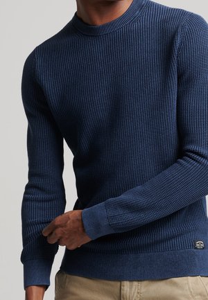 Homme portant un pull à manches longues bleu marine texturé ajustant le poignet, assorti à un pantalon beige.