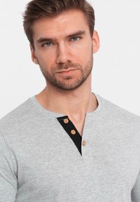 Grijs hemd met lange mouwen, voorzien van een zwarte knoopsluiting en houten knopen. Zachte textuur, casual ontwerp en een ronde halslijn.