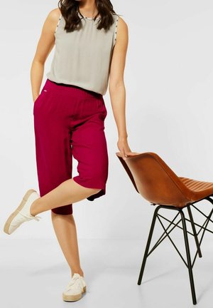 Pantaloni capri bordeaux con vita elasticizzata, abbinati a una maglietta senza maniche grigia chiara. Il modello si trova accanto a una sedia marrone. Indossa delle sneakers bianche.