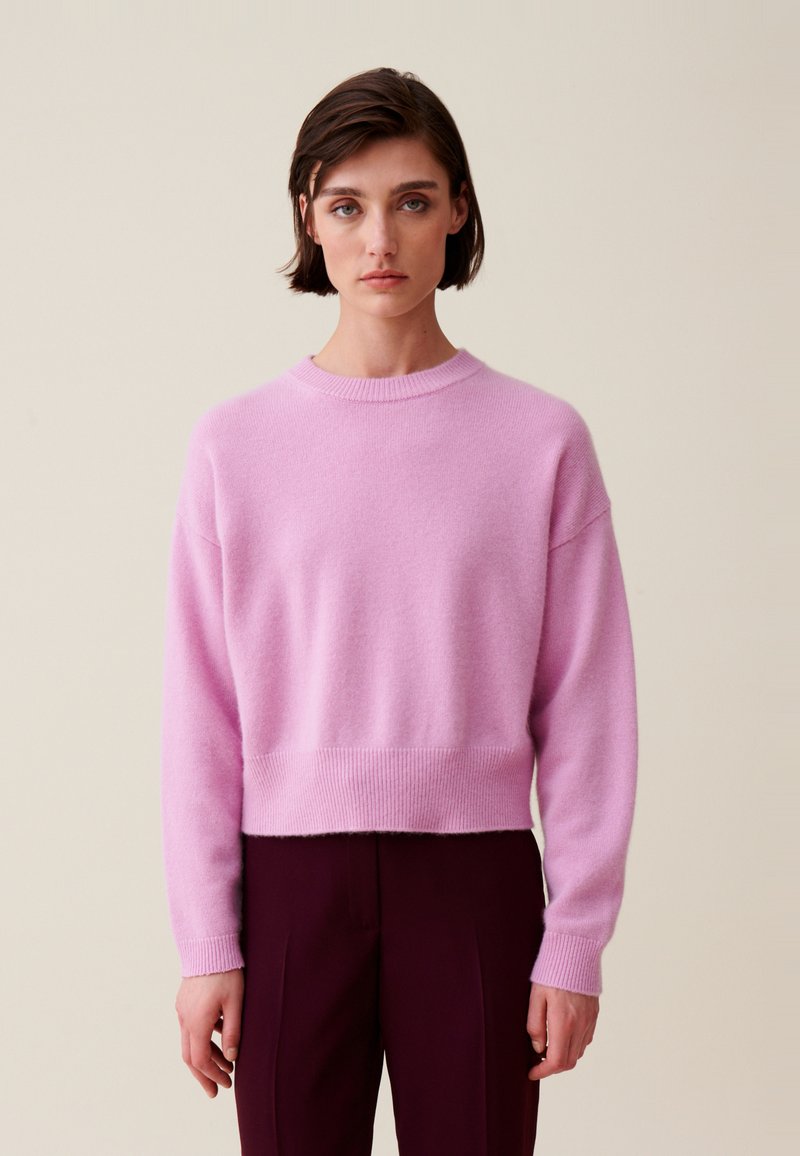 Pull court rose en tissu doux, avec un col rond, une coupe décontractée et des poignets et un ourlet côtelés. Porté avec un pantalon sombre.