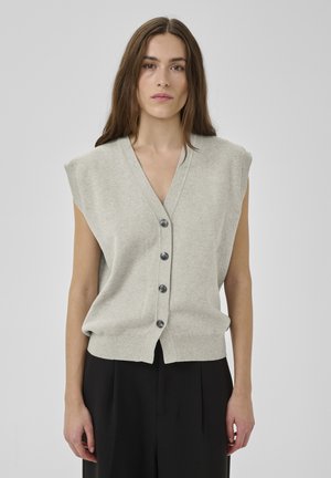 Moteris su ilgais rudais plaukais, vilkinti be rankovių šviesiai pilką liemenę su sagomis ir juodus kelnes, stovinti priešais paprastą šviesią foną.