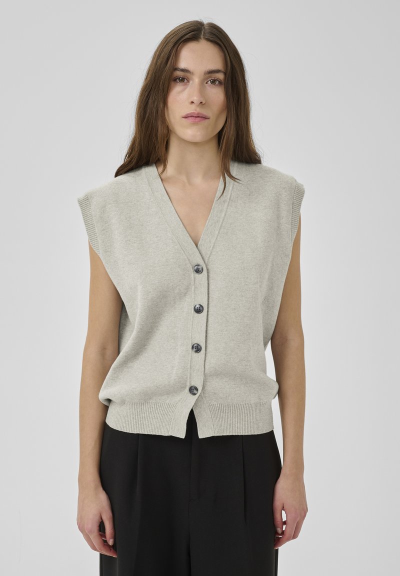 Pikkade pruunide juustega naine, kes kannab varrukateta helehallide nööbitud vestiga ja musti pükse, seisab ühevärvilise heleda tausta ees.