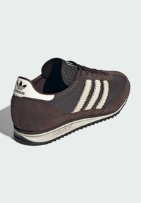 Smeđe i bež Adidas tenisice s tri pruge, vezanje sprijeda i teksturiranom potplatom, prikazane iz kuta straga na svijetloj pozadini.