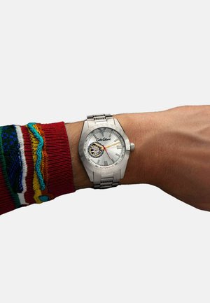 Silver automatic watch with a textured bezel and a white dial, featuring gold accents and a visible movement, worn on a wrist with a colorful sweater.
