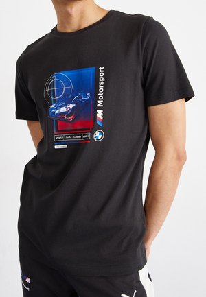 Osoba nosící černé tričko BMW Motorsport s grafikou modrého a červeného závodního vozu a textovými detaily na přední straně.