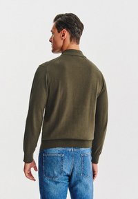 Zielony oliwkowy sweter z kołnierzykiem, ściągaczami przy mankietach i u dołu, w połączeniu z jasnoniebieskimi dżinsami, widok od tyłu.