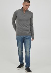 Maglione grigio con zip in una texture morbida, abbinato a jeans azzurri chiari e sneakers nere. L'outfit presenta un design casual con una forma aderente.