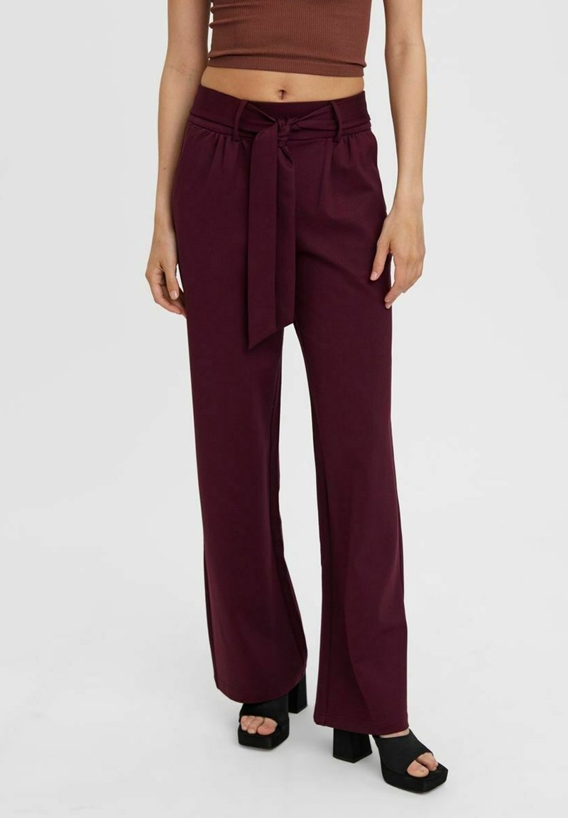 Femme portant un pantalon bordeaux taille haute à jambes larges avec une ceinture à nouer, associé à des sandales plateformes noires à bout ouvert et un top court marron.