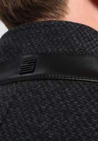 Gros plan sur le col d'un manteau texturé gris foncé avec des bordures en cuir noir et un petit logo métallique noir à l'arrière, porté par une personne aux cheveux bruns courts.