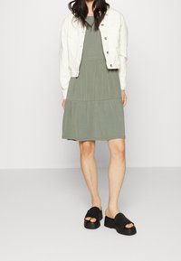 Olivově zelené vrstvené šaty s kulatým výstřihem, doplněné o zkrácenou bílou džínovou bundu a černé platformové slip-on boty.