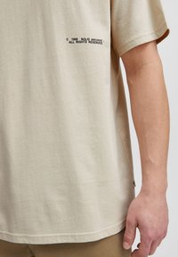 Beige kortærmet t-shirt lavet af bomuld. Har et lille sort tryk med tekst på siden. Blød tekstur, afslappet pasform og sideslids.