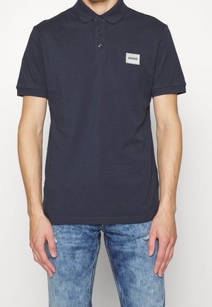 Mann trägt dunkelblaues Poloshirt mit kurzen Ärmeln und „HUGO“-Logoaufnäher auf der Brust, kombiniert mit hellblauen Jeans.