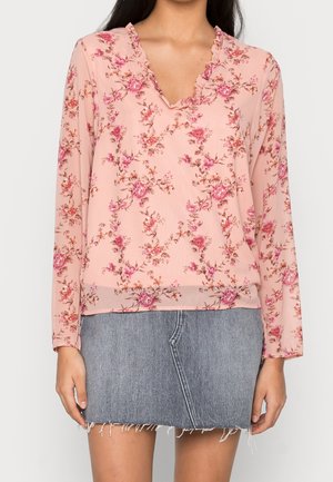 Kvinden har en pink langærmet bluse med blomstermønster og flæser ved V-udskæringen på, kombineret med en grå denim mininederdel med frynset kant.