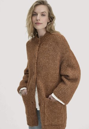 Frau in einer grob gestrickten braunen Strickjacke mit großen Taschen vorne, die über einem weißen Shirt und blauen Jeans steht, vor einem schlichten Hintergrund.