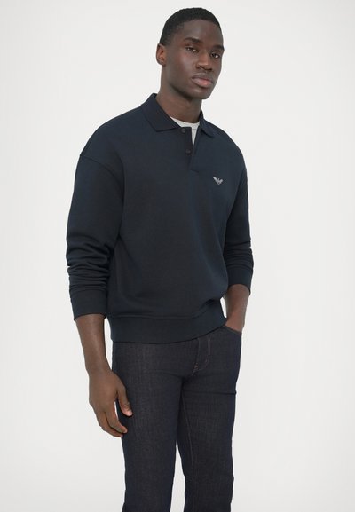 Tmavomodrá polokošelárová mikina z bavlneného zmesi s okrúhlym výstrihom, dlhými rukávmi a jemným logom na hrudi. Outfit dopĺňajú riflové nohavice.