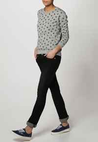 Felpa grigia con piccoli motivi neri, abbinata a jeans neri aderenti e sneakers blu scuro. Design casual con polsini a coste.