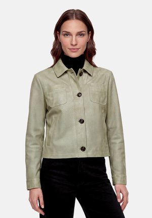 Femme portant une veste en cuir vert clair boutonnée par-dessus un col roulé noir et un pantalon noir, debout devant un fond uni.