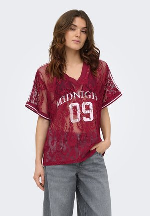 Femeie purtând o cămașă din dantelă burgundy transparentă cu textul "MIDNIGHT 09" și blugi bleu deschis, stând cu o mână în buzunar.