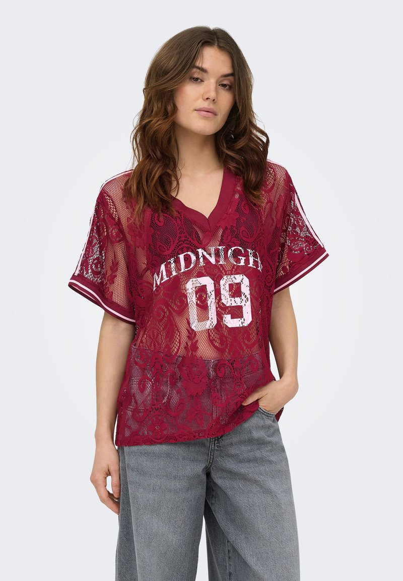 Femme portant une chemise en dentelle bordeaux transparente avec le texte "MIDNIGHT 09" et un jean bleu clair, debout avec une main dans la poche.