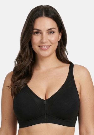 Smilende kvinde med langt mørkt hår iført en sort front-lynlås bh, vendt mod kameraet foran en ensartet lys baggrund.