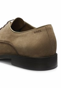 Chaussure habillée en daim marron avec semelle foncée, bout rond, lacets, et "BOSS" embossé près du talon sur le côté extérieur.