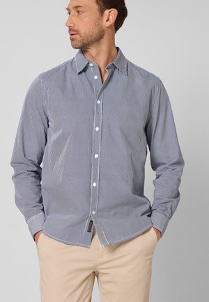 Mann trägt blau-weiß gestreiftes, dünnes Hemd mit Knöpfen, beige Hose, Hände in den Taschen, schaut nach links vor schlichtem grauem Hintergrund.