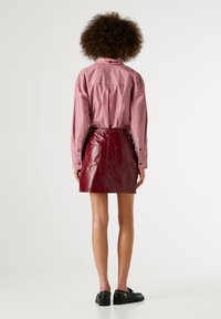Mini-jupe en cuir verni rouge à taille haute et poches arrière, portée avec une chemise à manches longues rayée rose et blanche. Chaussures noires.