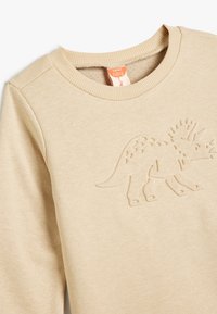 Suéter de manga larga beige con un diseño texturizado y en relieve de un triceratops en la parte frontal. Presenta un cuello redondo acanalado. Material de algodón suave.