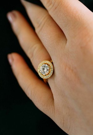 Goldring mit einem großen zentralen Diamanten, umgeben von kleineren Diamanten, am Ringfinger einer Hand vor einem dunklen Hintergrund.