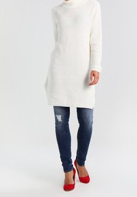 Weißes Strick-Turtleneck-Kleid mit lockerem Schnitt, kombiniert mit verwaschenen Blue Jeans und roten High Heels. Strukturierter Stoff sichtbar.