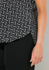 Blusa a zigzag bianca e nera con maniche corte, spacco laterale e texture liscia, abbinata a pantaloni neri.