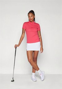 Rózsaszín galléros piké ing, öt gombos sliccel, fehér golf szoknyával párosítva. A modell egy golfütőt tart, és fehér tornacipőt visel.